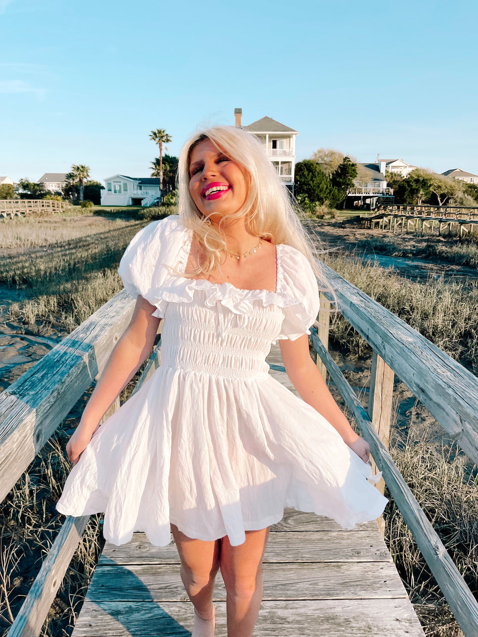 White mini dress with puff sleeves and smocked bodice, worn outdoors near water.
