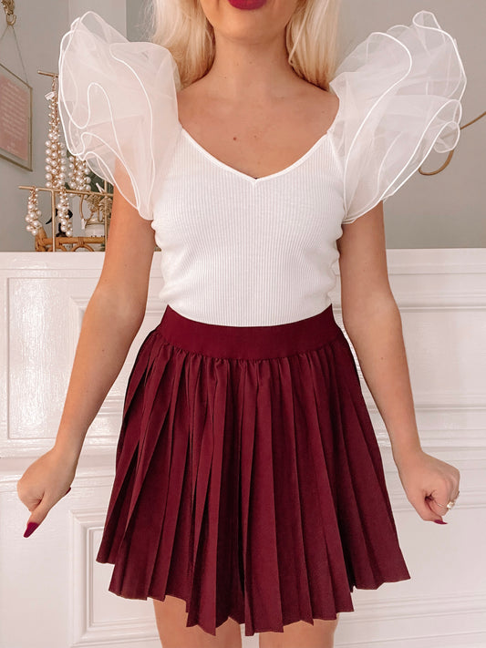 Burgundy pleated mini skirt paired with a white ribbed top featuring sheer, ruffled sleeves.
