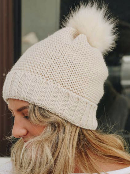 Cream knit beanie with fluffy pompom, worn by a blonde woman.
