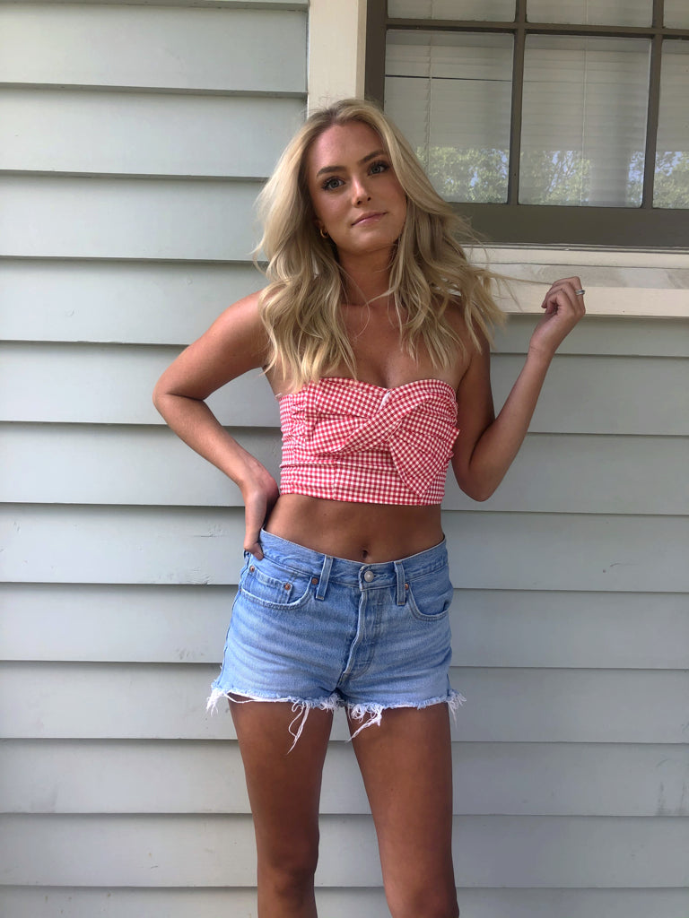 Woman in red gingham crop top and denim shorts against light gray siding.
